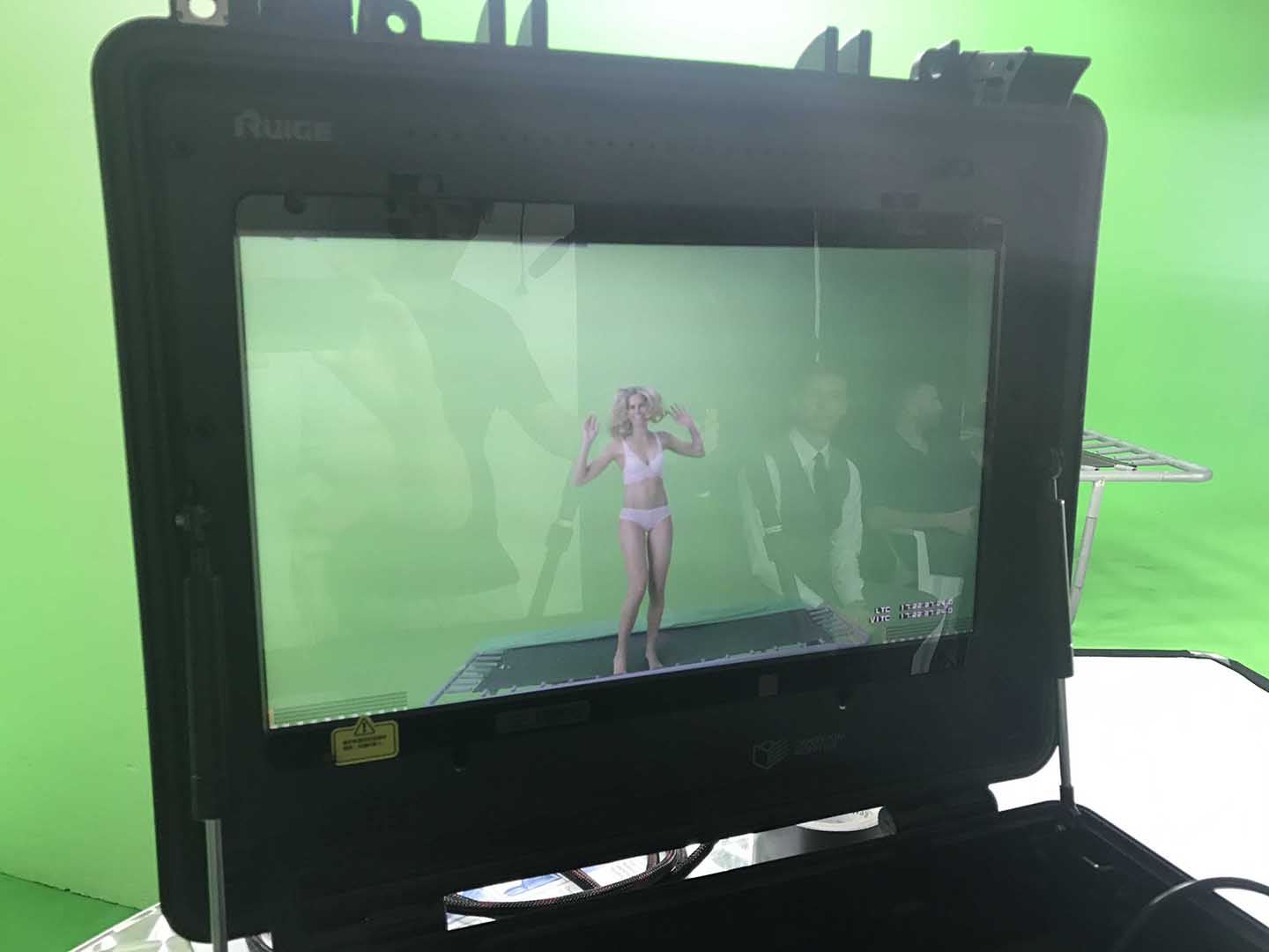 Through a monitor we see a female model on a trampoline in a green screen studio.