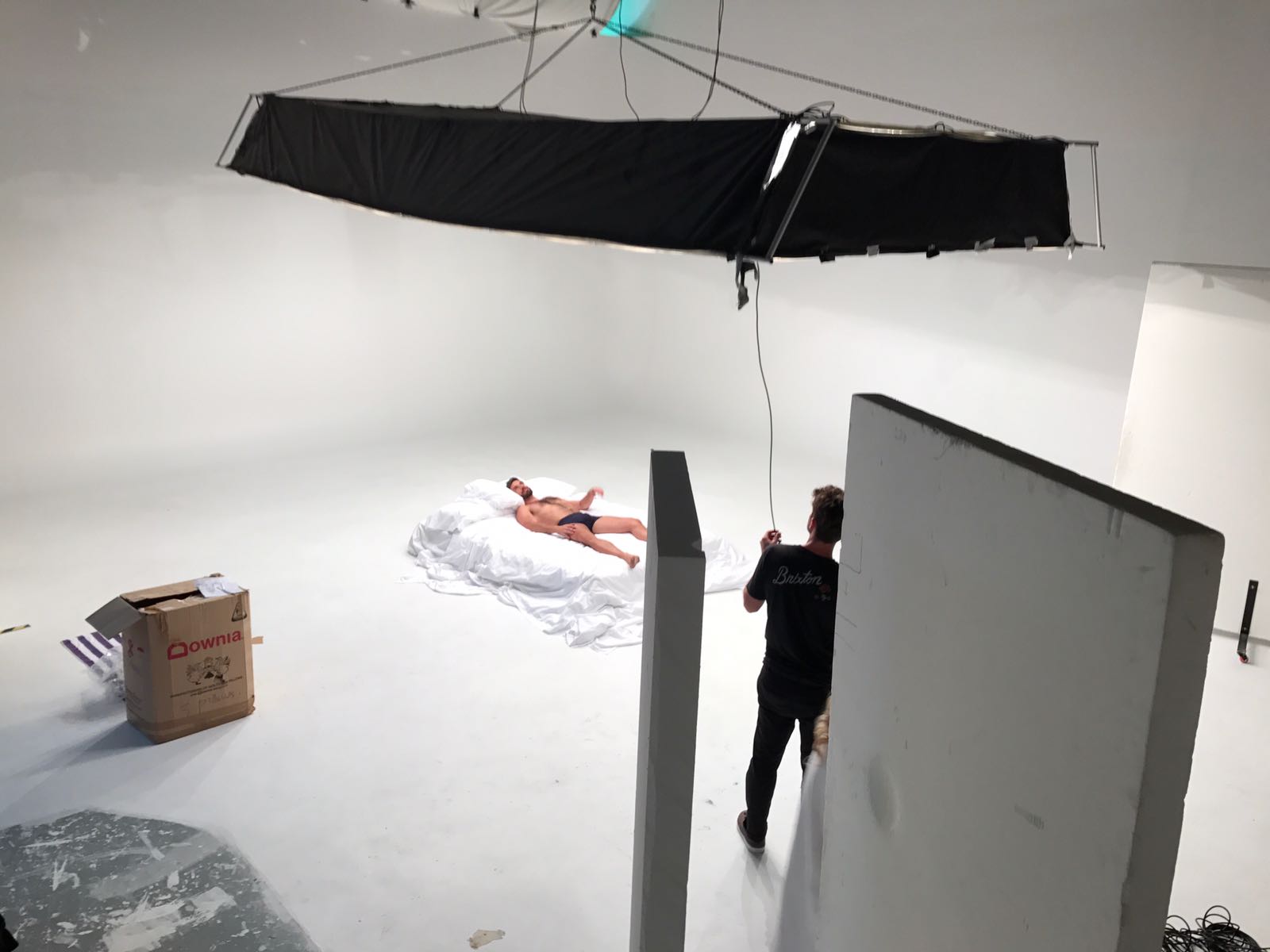 A male model lies on a white bed while being filmed in a studio.